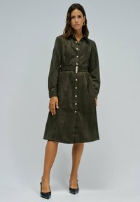 Robe chemise vert olive à manches longues avec une boutonnière sur le devant, une taille cintrée et une boucle de ceinture argentée brillante. Texture lisse semblable au daim.