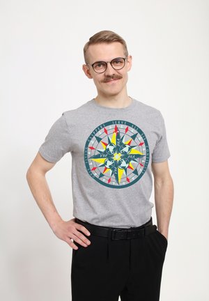 Grå kortærmet T-shirt med et cirkulært kompasdesign i blå, gul og rød; parret med sorte bukser.