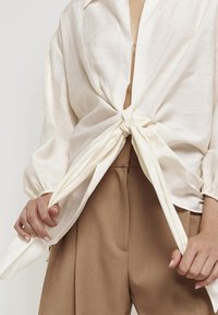 White, lightweight, long-sleeve top with a knotted front, worn over high-waisted brown trousers. Smooth texture, relaxed fit, and soft sheen.