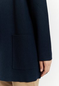 Maglione blu navy in tessuto di lana testurizzato, con una tasca laterale e un orlo leggermente curvo, parzialmente coperto da una mano.