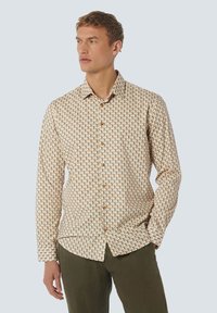 Man in een beige overhemd met knopen en een geometrisch patroon, met olijfgroene broek, kijkend opzij met één hand in de zak.