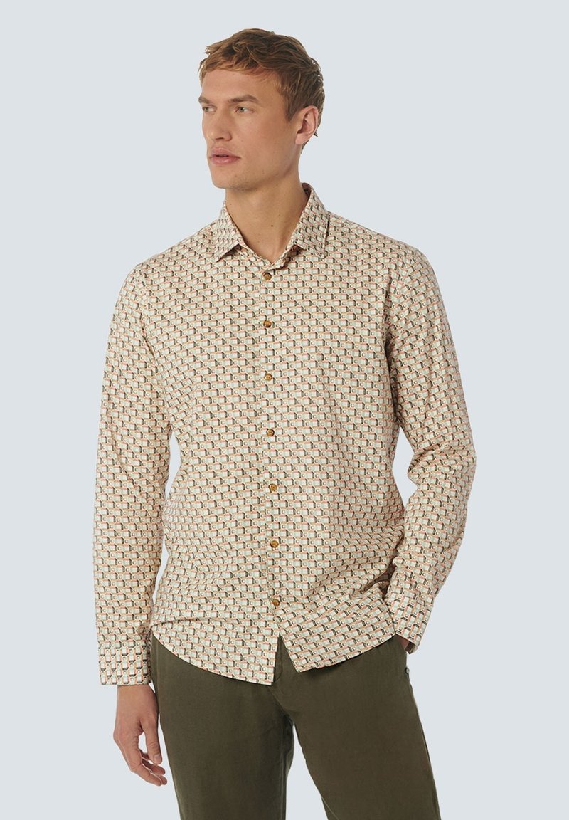 Man in een beige overhemd met knopen en een geometrisch patroon, met olijfgroene broek, kijkend opzij met één hand in de zak.