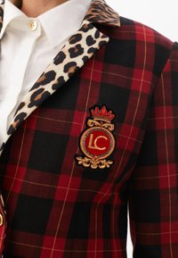 Dettaglio di un blazer con un motivo scozzese rosso e nero, colletto in stampa leopardata e un distintivo decorativo con "LC" e un emblema di corona.