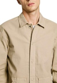 Giacca beige con bottoni, dotata di colletto, caratterizzata da un design minimalista e due tasche frontali realizzate in un tessuto testurizzato.