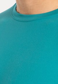 Camisa corta de manga, color turquesa, con cuello redondo, hecha de una tela suave y flexible. Presenta un diseño simple y sin costuras, sin patrones.