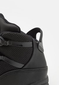 Svart sneaker med en texturerad mesh-överdel, släta läderdetaljer och en dragflik. Synlig snörning och en strömlinjeformad design.