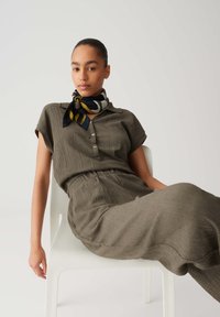 Olive-green, pleated dress with short sleeves, button-down front, elastic waist, and a patterned scarf in navy, yellow, and white at the neck.