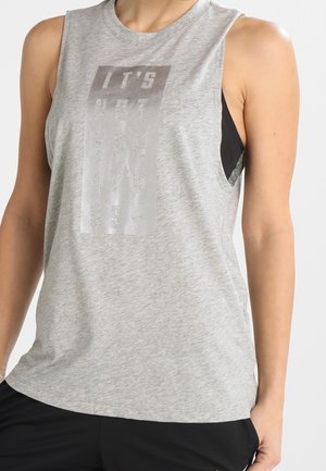 Haut de sport sans manches gris clair avec un soutien-gorge de sport noir visible en dessous, porté par une personne avec la main droite dans la poche d’un short noir.