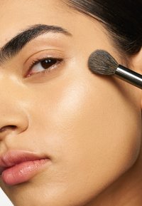 Pinceau de maquillage appliquant de la poudre sur une peau lisse et radieuse, mettant en valeur un teint naturel avec une légère brillance et un accent sur des sourcils et des lèvres bien définis.