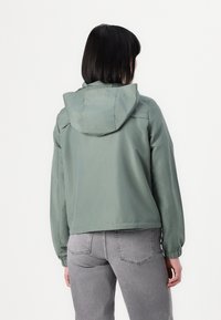Light green hooded jacket with elastic cuffs and a cropped design, made from smooth, water-resistant fabric. Back view highlights stitching details.