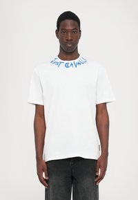 Hvit bomullst-skjorte med korte ermer og rund hals, med blå brodert tekst rundt kragen. Kombinert med mørke jeans.