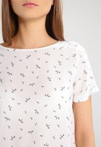 Hvid kortærmet bluse med sorte guldsmedemønstre. Letvægtsstof, rund halsudskæring og detaljer med opsmøgede ærmekanter. Blødt tekstur.