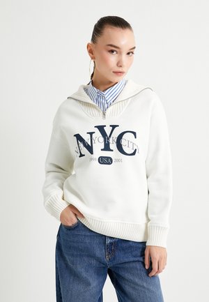 Felpa bianca con grafica "NYC", orlo e polsini a costine, colletto a zip lungo un quarto, indossata sopra una camicia a righe blu e bianca, abbinata a jeans di denim.