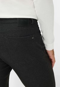 Pantalon en denim noir avec une finition texturée, présentant une poche arrière et un patch en cuir avec logo. La taille est dotée de passants de ceinture et d'un design épuré.