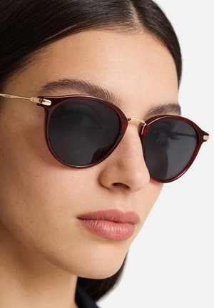 Occhiali da sole rotondi con montatura color bordeaux, lenti scure e dettagli dorati. Sottili aste che si estendono dalle lenti alle orecchie. Texture liscia.