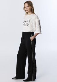 Top bianco testurizzato con la scritta "MERCI AMOUR" in lettere nere, abbinato a pantaloni neri a vita alta con strisce laterali bianche. Scarpe nere.