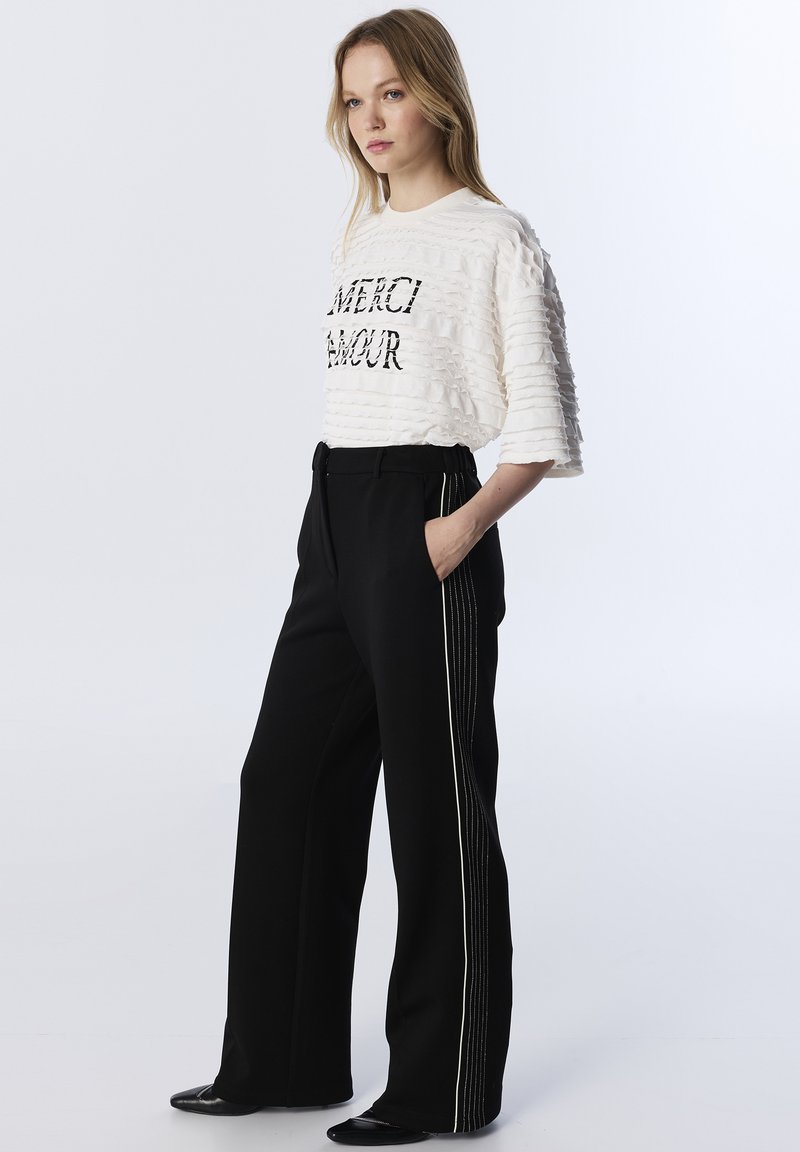 Top bianco testurizzato con la scritta "MERCI AMOUR" in lettere nere, abbinato a pantaloni neri a vita alta con strisce laterali bianche. Scarpe nere.