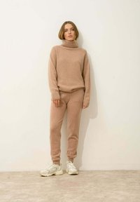 Jasno beżowy sweter z dzianiny z wysokim golfem i luźnym krojem, w połączeniu z dopasowanymi spodniami dresowymi. Białe sneakersy dopełniają stylizację.