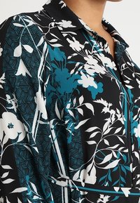 Blus med blommönster på svart bakgrund, med vita och turkosa mönster, kragdesign och en slät, lätt tygtextur.