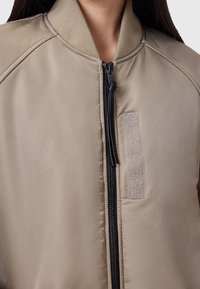 Helles beige Bomberjacke mit Reißverschluss vorne, geripptem Kragen und strukturierter Stoff mit Velcro-Patch-Detail.