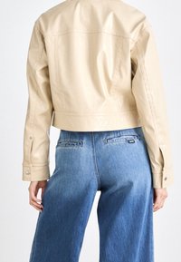 Beżowa kurtka z syntetycznej skóry o kroju crop, gładkiej fakturze, z zapięciem na guziki przy mankietach oraz prostym designie, zestawiona z niebieskimi dżinsami o wysokim stanem.