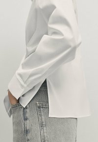 Hvid langærmet skjorte med en glat tekstur, der har en delt kant og knapmanchetter. Sammen med lyseblå denimjeans.