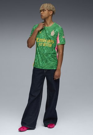 Maglia da calcio verde con motivi a foglia, dettagli rosa e testo giallo. Abbinata a jeans larghi blu scuro e calzature rosa.