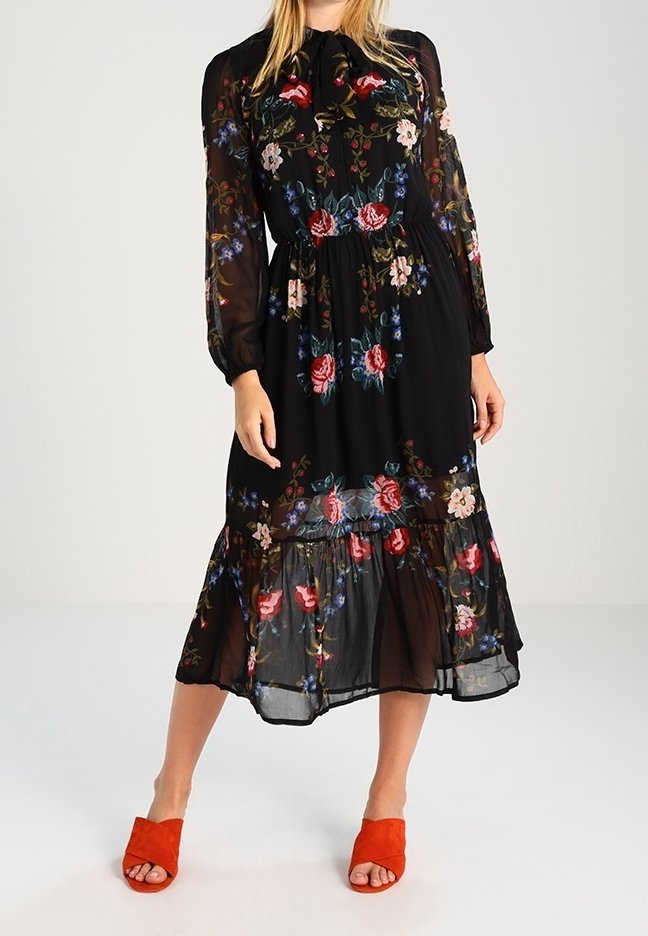 Femme portant une robe midi noire transparente avec une broderie florale multicolore et des manches longues, associée à des talons ouverts orange vif.