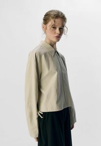 Giacca beige con zip frontale, colletto e maniche lunghe. Presenta dettagli con coulisse elastica all'orlo per una vestibilità regolabile. Tessuto morbido.