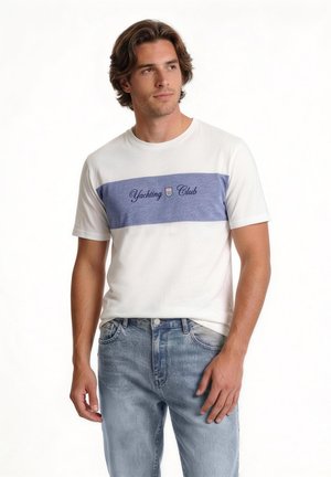 Młody mężczyzna w białej i niebieskiej koszulce "Yachting Club" oraz jasnoniebieskich dżinsach, stoi i patrzy na bok na białym tle.