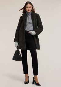 Tmavě šedý, texturovaný shag kabát přes šedý rolák, doplněný černými zúženými kalhotami a lakovanými lodičkami. Drží černou kabelku.