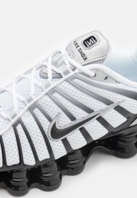 Hvit Nike Shox-sneaker med mesh-overdel, svarte detaljer, sølvlogo og distinkt polstret såle med rundede pigger.