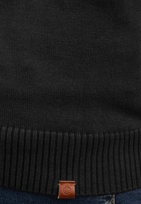 Schwarzer Strickpullover mit geripptem Saum und einem braunen Lederetikett am unteren Ende, vor einem Denim-Hintergrund präsentiert.