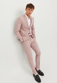 Costume ajusté rose clair, coupe slim, veste à un bouton, pantalon à devant plat, associé à une chemise blanche et des mocassins noirs.