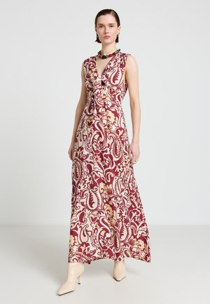 Donna che indossa un abito senza maniche lungo fino ai piedi con motivo paisley floreale rosso e beige, accessoriato con una collana spessa e stivali chiari.