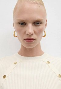 Detailní záběr osoby s hladce učesanými blond vlasy, modrýma očima, která má na sobě zlaté kroužky a bílý žebrovaný top se zlatými knoflíky.