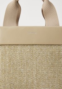Bežna tkanina torba s glatkim bež usnjenim vrhom in kontrastnimi teksturiranimi naramnicami. Na sprednji strani je vrezan logotip "Calvin Klein".
