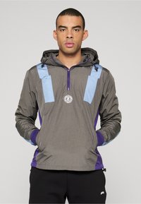 Graue leichte Jacke mit blauen Akzenten und lila Details, mit Reißverschluss vorne und Kapuze, strukturiertem Stoff und rundem Logo auf der Brust.