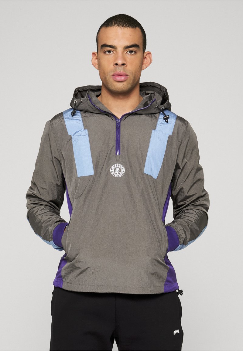 Graue leichte Jacke mit blauen Akzenten und lila Details, mit Reißverschluss vorne und Kapuze, strukturiertem Stoff und rundem Logo auf der Brust.