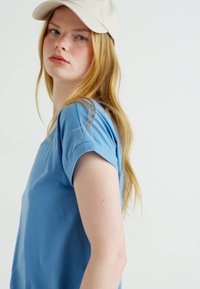 T-shirt à manches courtes bleu clair en tissu doux, avec des manches retroussées. Le modèle porte une casquette de baseball beige, légèrement de côté.