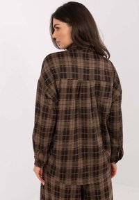 Plaid shirt in brown and tan tones, featuring a high collar, loose fit, and button accents on the sleeves. Textured, soft fabric.
