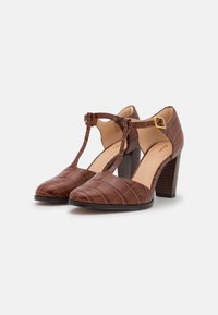 Escarpins T-strap marron avec motif crocodile texturé, détail de boucle à la cheville et talon bloc empilé. Doublure intérieure lisse.