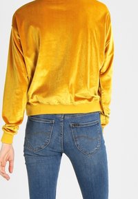 Top amarillo de terciopelo de manga larga con puños y dobladillo acanalados, combinado con jeans de mezclilla azul que tienen bolsillos traseros y un ligero desgaste.