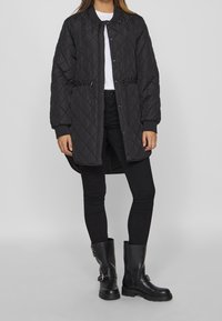 Vadderad svart jacka med rak skärning, elastisk midja och tryckknappsstängning. Kombinerad med svarta slim-fit jeans och svarta stövlar med spänne.