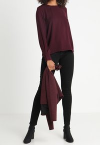 Blouse bordeaux à manches longues avec un col rond, associée à un jean skinny noir et des bottines, tenant un gilet assorti.