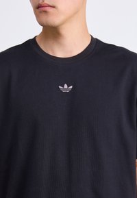 Vyras dėvi paprastą juodą apvalios kaklo marškinėlį su nedideliu siuvinėtu Adidas logotipu, kuris yra centre ant krūtinės.
