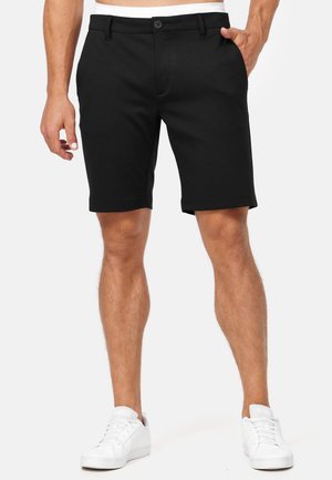 Männlich, der schwarze geknöpfte Shorts mit Seitentaschen und weißen Sneakers trägt, steht mit einer Hand in der Tasche vor weißem Hintergrund.