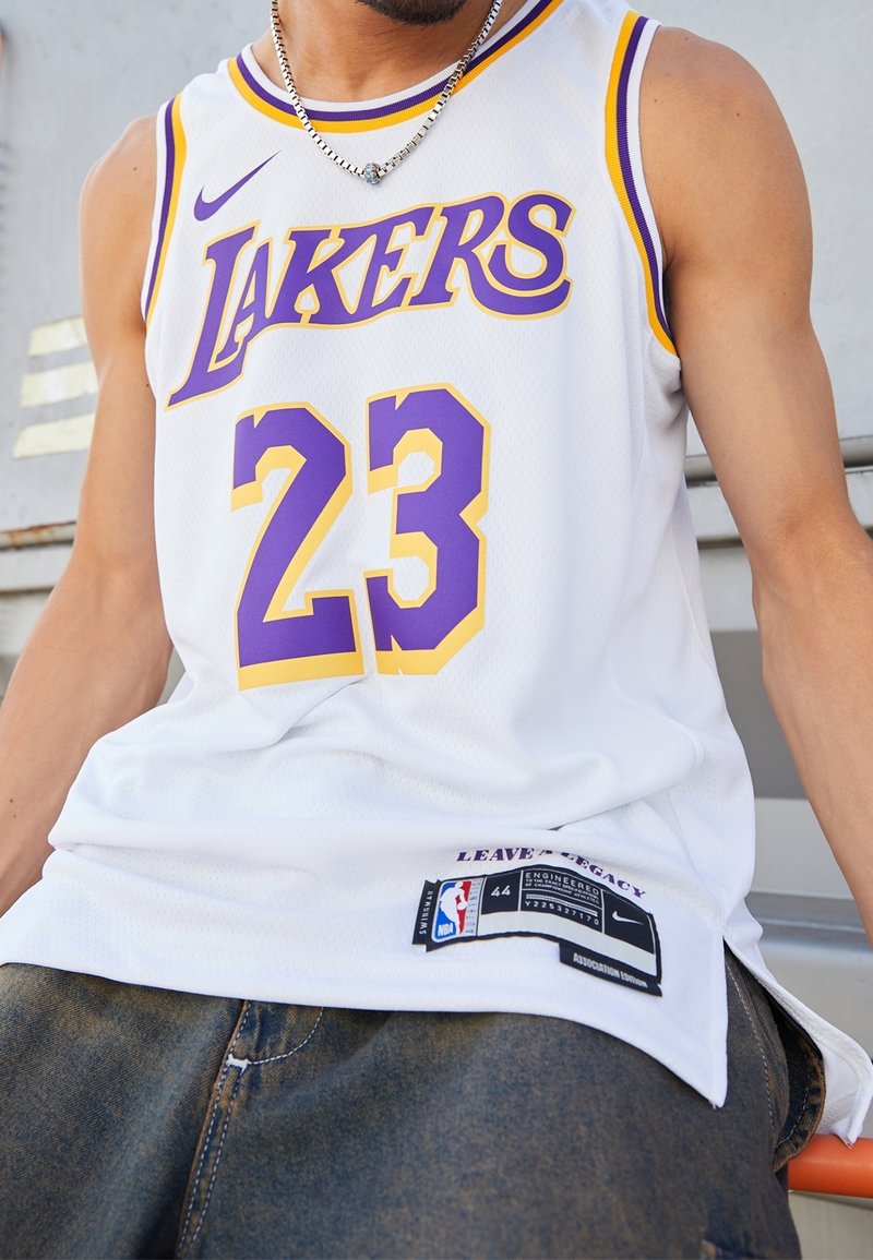 Weißes Basketball-Trikot mit lila und gelben Akzenten, ausgestattet mit "LAKERS" und der Nummer "23". Nike-Logo und NBA-Tag am Saum. Netzstruktur.