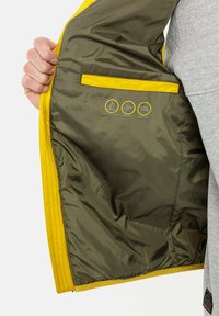 Gul jakke med olivengrønt for; har lommer og ikoner, der angiver vandafvisende, vindtæt og dunfri egenskaber.