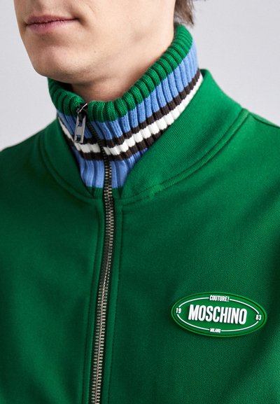 Zelená zipová bunda s ribovanými akcentmi v modrej, čiernej a bielej farbe na golieri a s označením loga na hrudi. Hladká textúra látky.